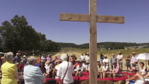 Scene from Half-Mile film shows wooden cross with folks sitting and gathered around in a rural setting